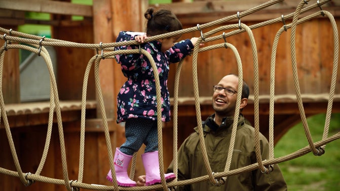 A young child walks along a rope bridge in the wild play area at Gibside while a parent smiles as he watches her.